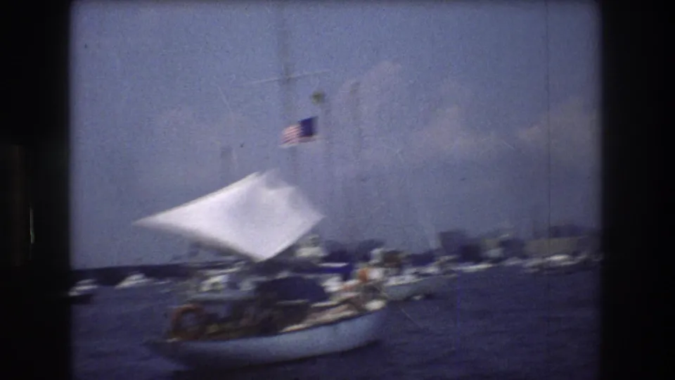 1976 New York City harbor home-movie frame showing a small sailboat with a white sail and American flag moving past other boats on blue water.