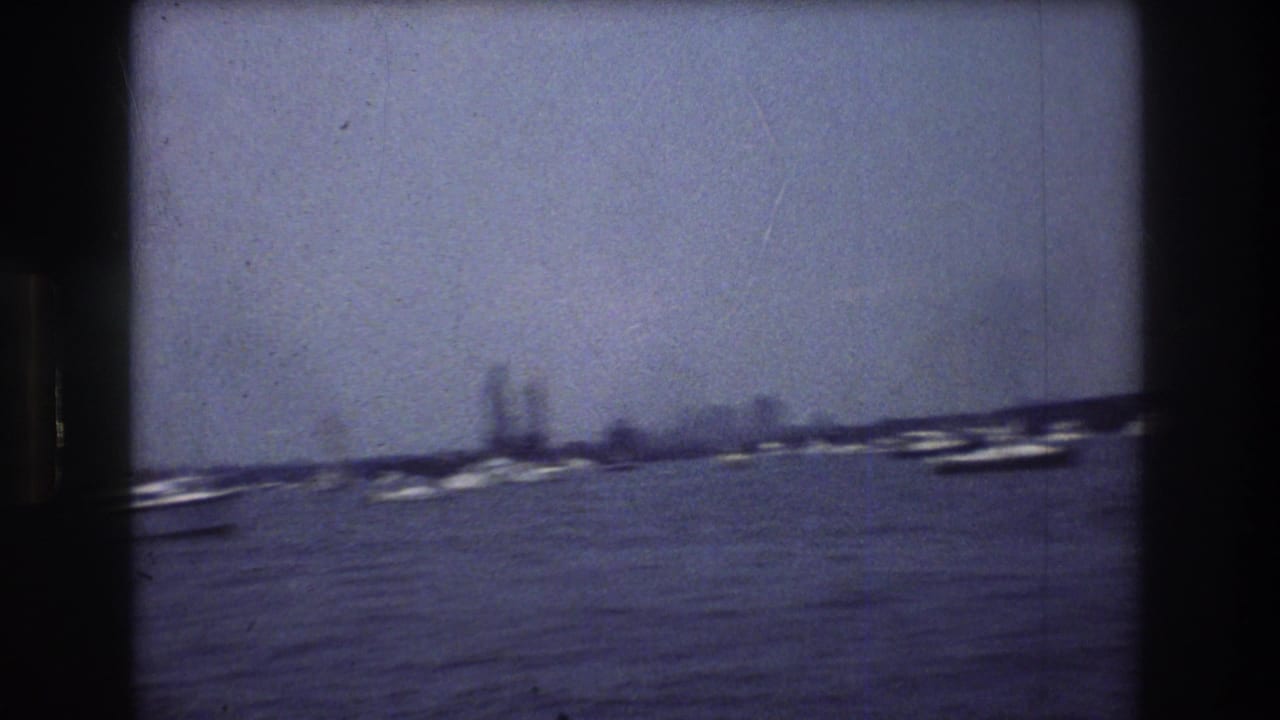 Soft 8mm frame of New York City harbor water, scattered boats, and a hazy distant shoreline with tall buildings visible in the background.