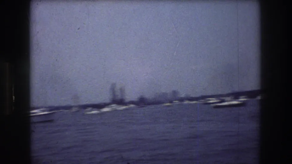 Soft 8mm frame of New York City harbor water, scattered boats, and a hazy distant shoreline with tall buildings visible in the background.