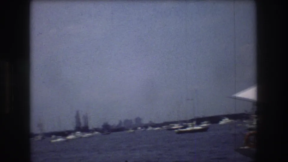 1976 New York City harbor frame showing open water, distant boats and buildings, and the edge of a white sail entering the right side of the frame.