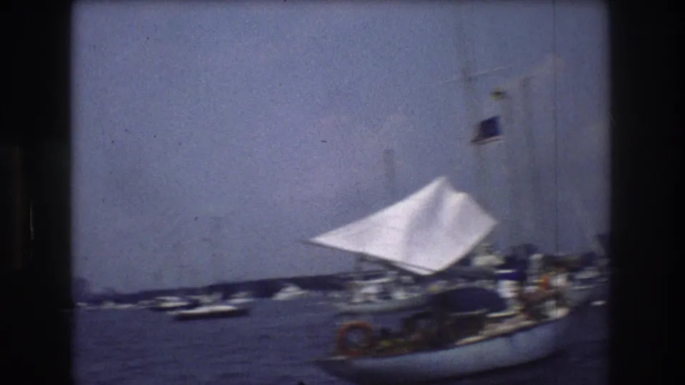 1976 8mm view of small boats and a sailboat on New York Harbor, with a hazy Manhattan skyline and a U.S. flag visible on one vessel.