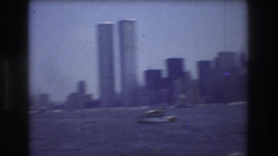 The Twin Towers and lower Manhattan skyline appear across New York Harbor, with small boats moving through the foreground water in the 1976 home-movie view.