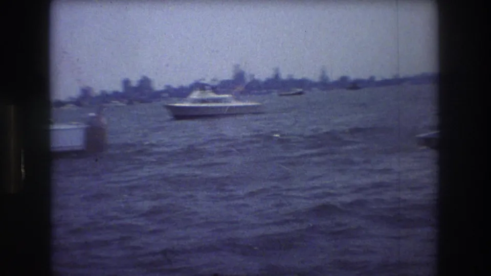 A white motorboat crosses New York Harbor in front of the distant New York City skyline, captured with soft focus and visible 8mm grain.