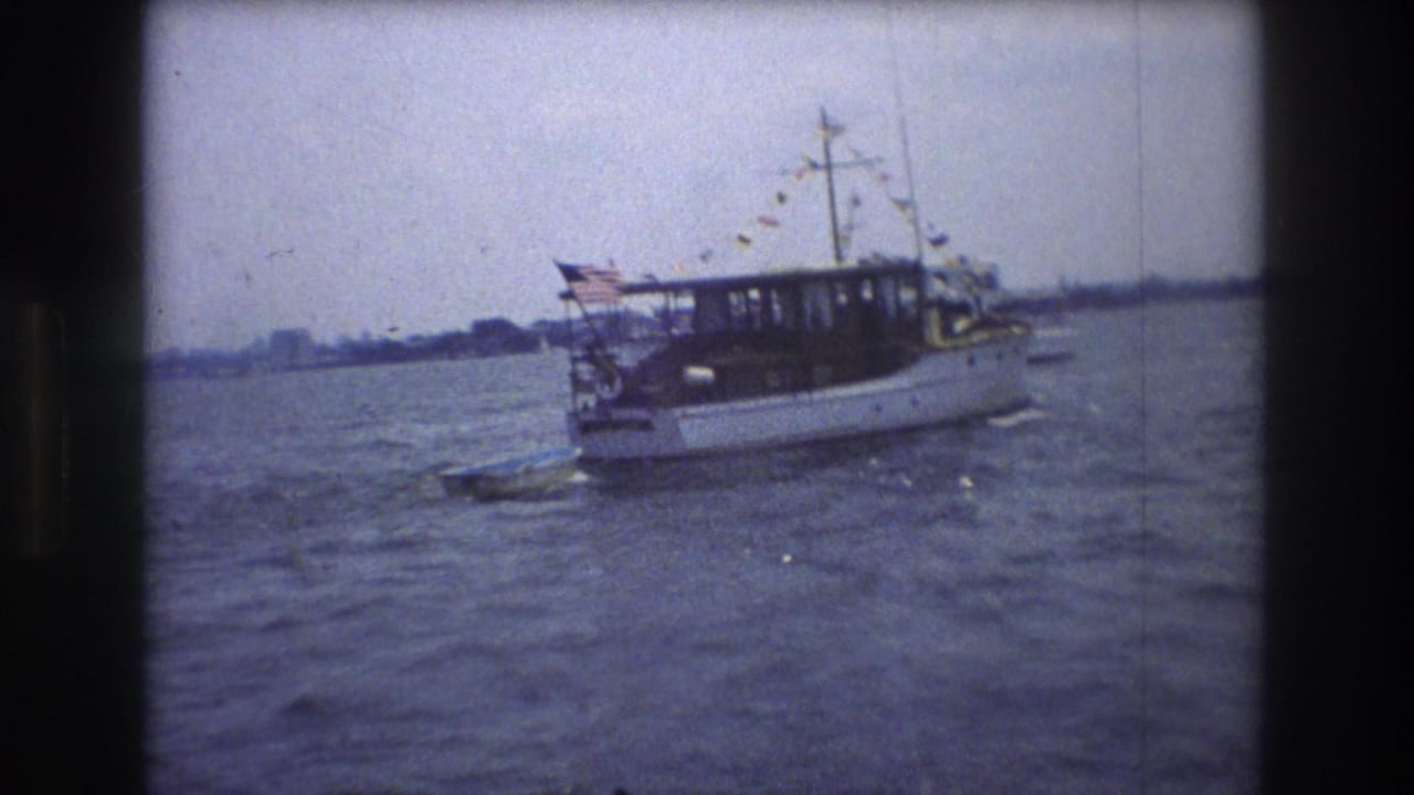 1976 New York City 8mm frame with a white motorboat carrying an American flag and string bunting on rippled water, with shoreline in the distance.
