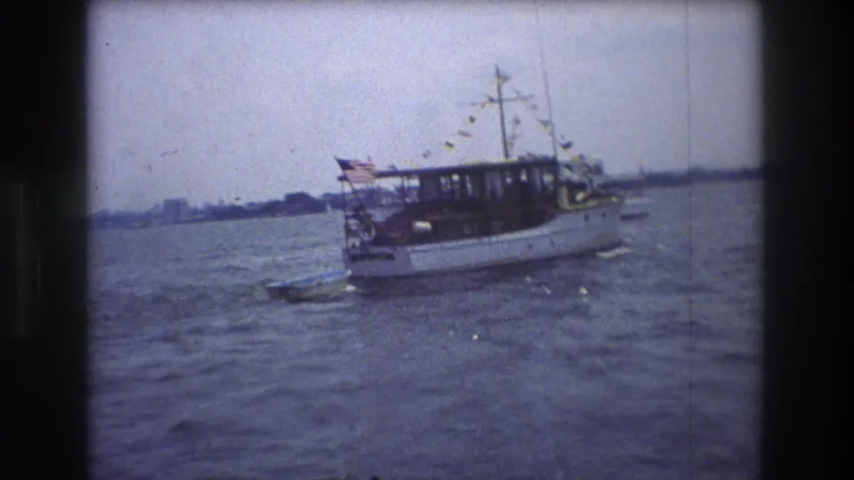 1976 New York City 8mm frame with a white motorboat carrying an American flag and string bunting on rippled water, with shoreline in the distance.
