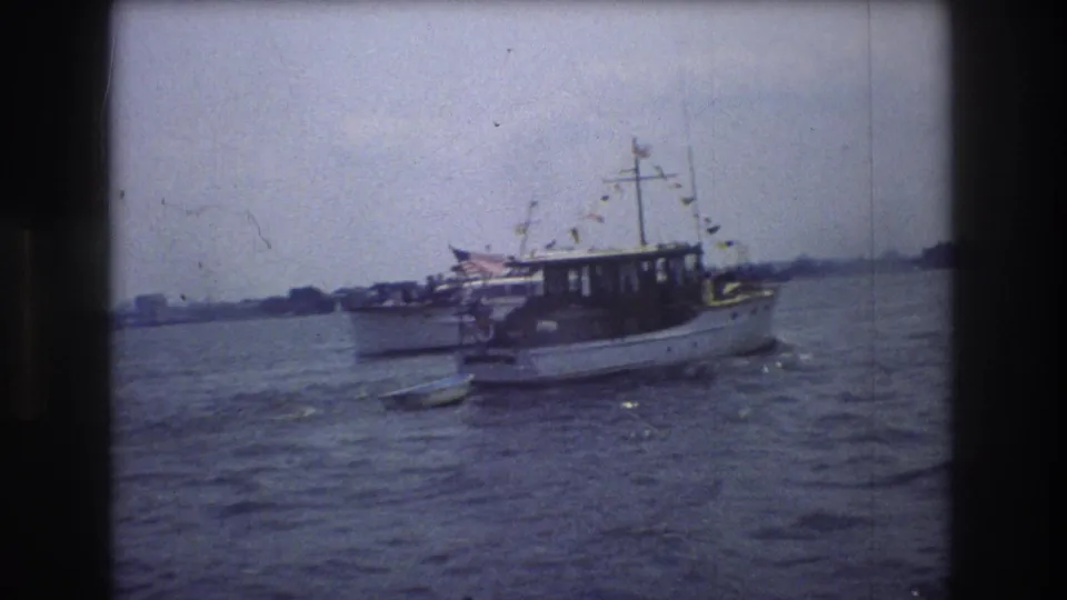 1976 New York City 8mm frame showing a decorated white motorboat centered on the water, with a small trailing object and hazy shoreline behind it.