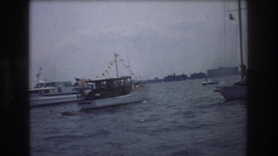 1976 New York City harbor footage showing a white motorboat with an American flag and bunting moving across choppy water near other boats.