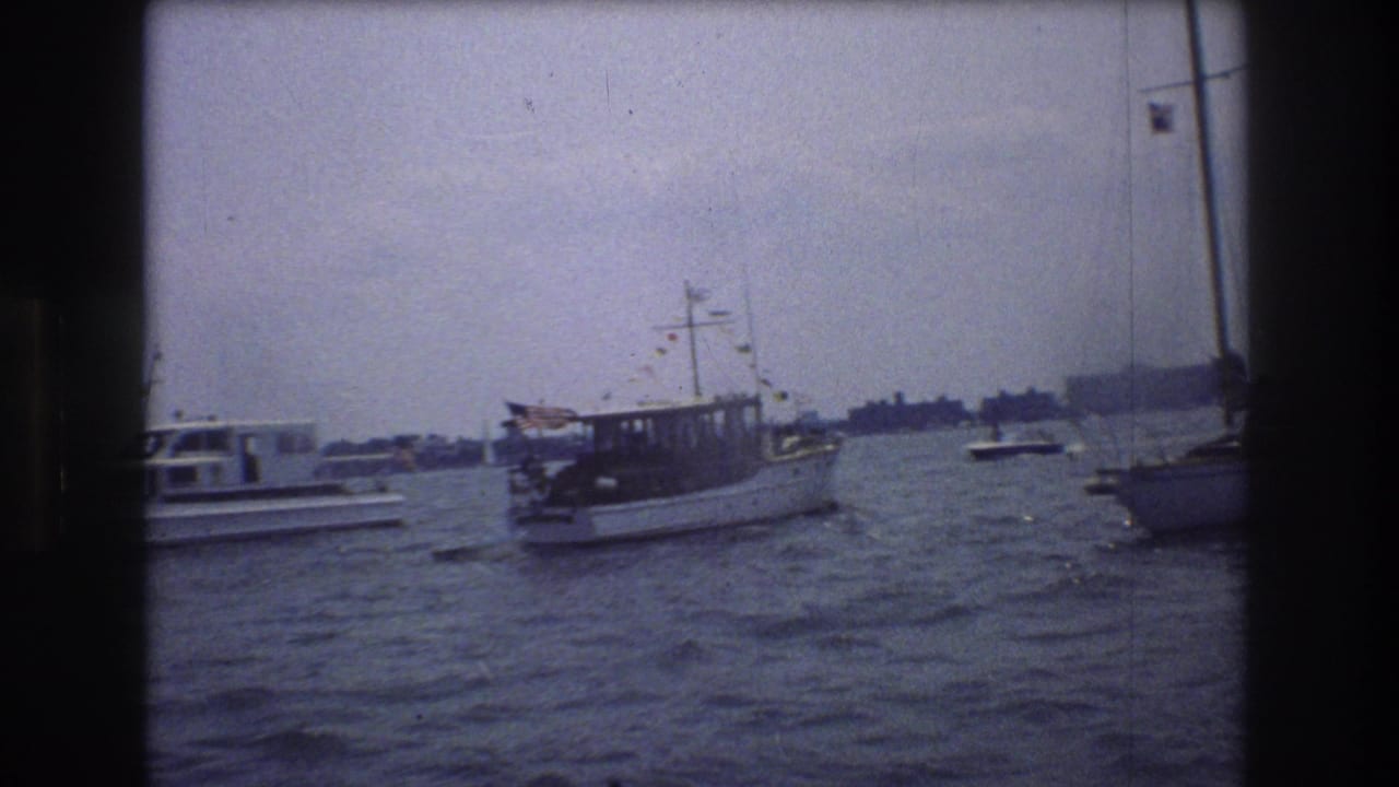 A small decorated motorboat appears in New York City harbor, with rough water, nearby vessels, and a low distant shoreline under a cloudy sky.