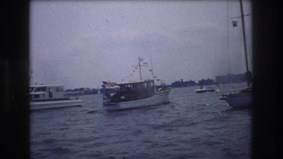 A small decorated motorboat appears in New York City harbor, with rough water, nearby vessels, and a low distant shoreline under a cloudy sky.