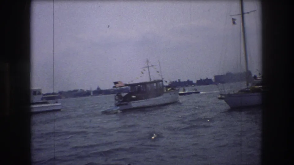 The handheld 8mm view holds on a flag-decorated motorboat moving through choppy New York City harbor water with other vessels nearby.