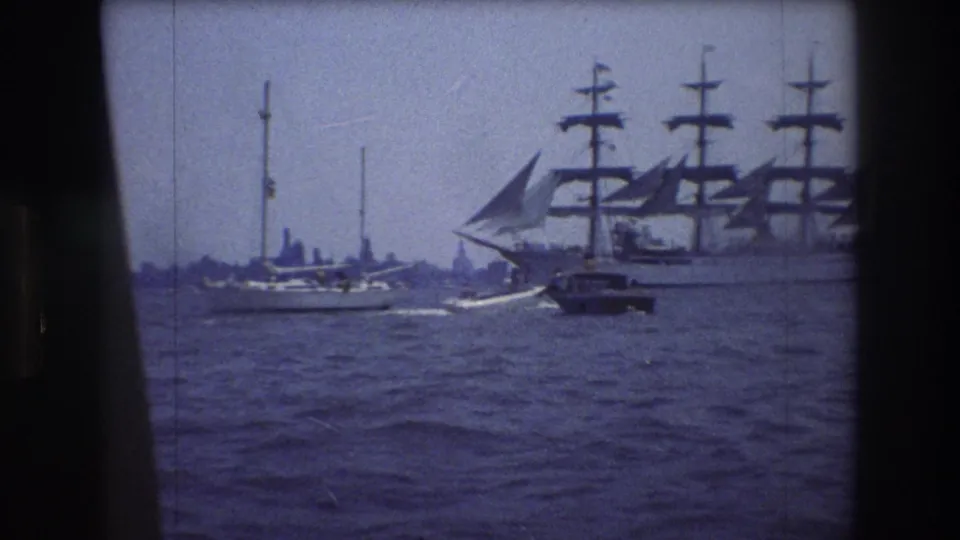 1976 New York City harbor view with a large multi-masted sailing ship, a small motorboat, and sailboats seen through an 8mm film frame.