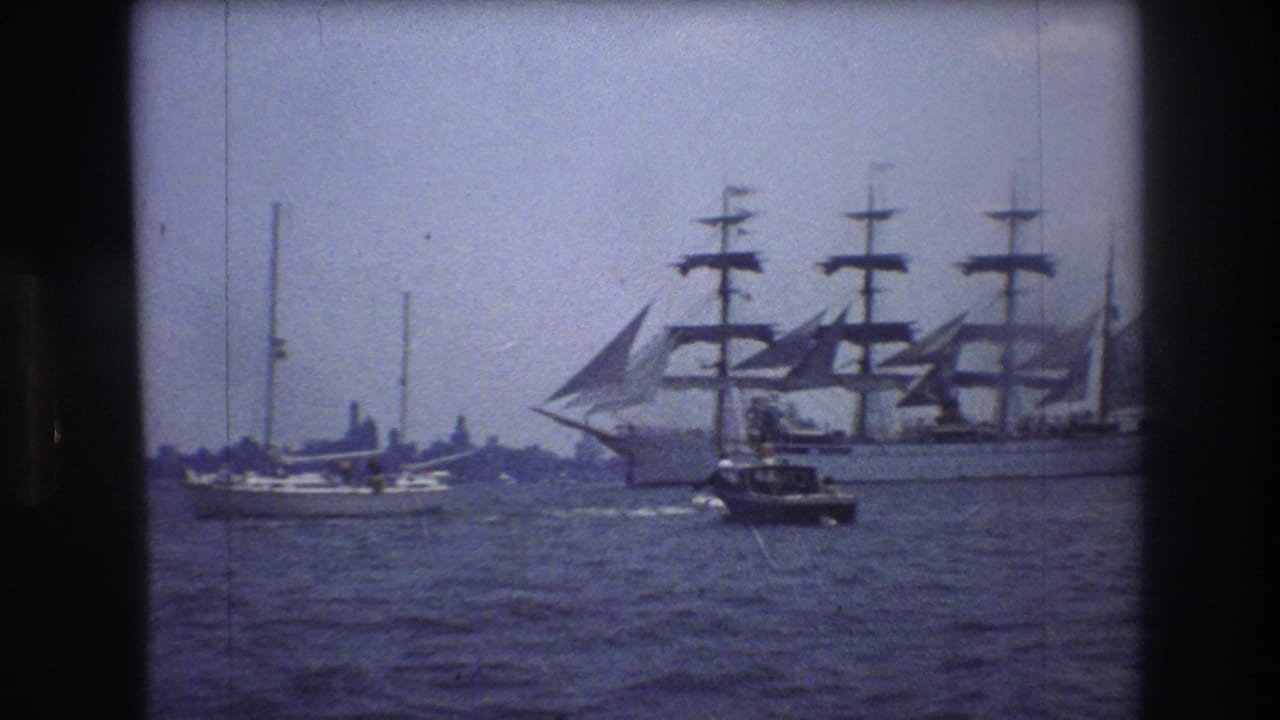 Tall sailing ship and nearby boats crossing a harbor under a pale sky.