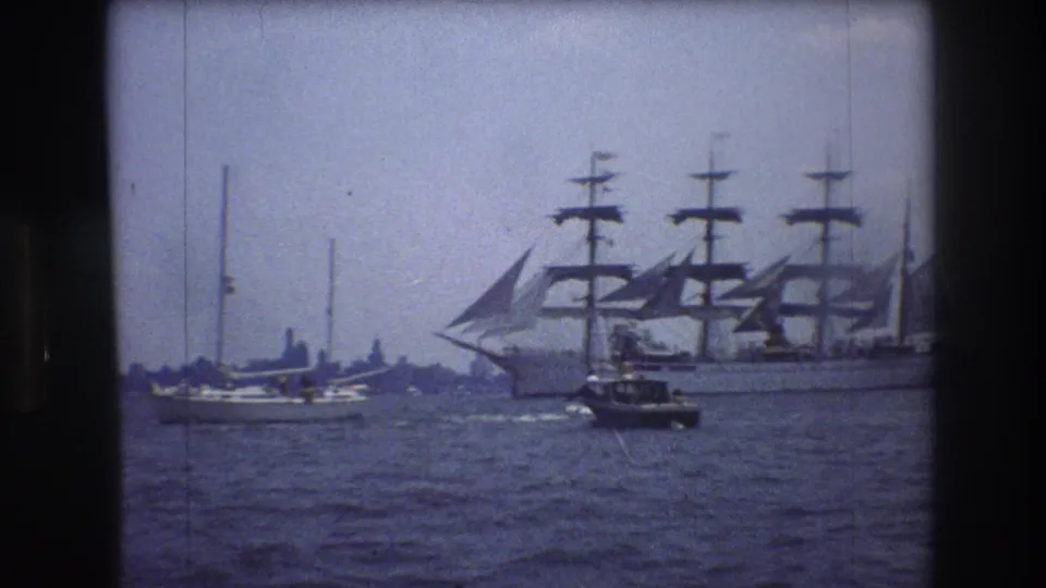 Large sailing ship with several masts moves through New York City harbor in 1976, with small boats nearby and a faint shoreline beyond.