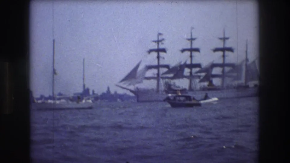 Home-movie frame from 1976 showing a multi-masted sailing ship in New York City harbor with a motorboat and sailboats crossing the foreground.