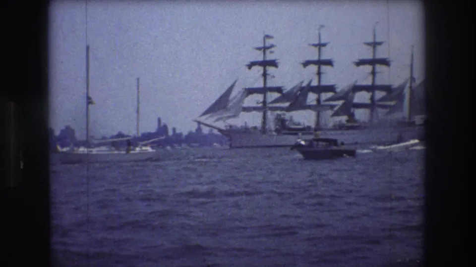 1976 New York City harbor footage frame with a tall sailing ship on the right, smaller boats in the foreground, and a hazy shoreline behind.