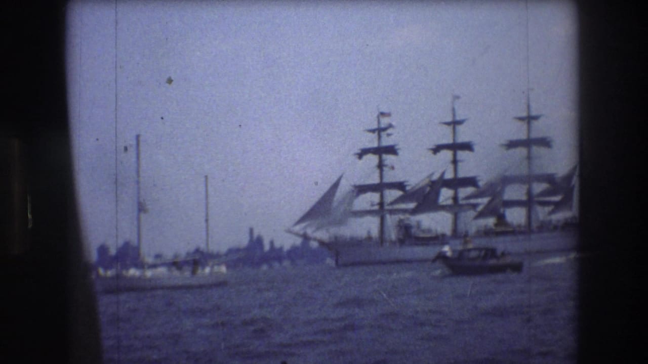 A large multi-masted sailing ship moves across New York City harbor water in 1976 home-movie footage, with smaller boats nearby and a hazy shoreline in the distance.