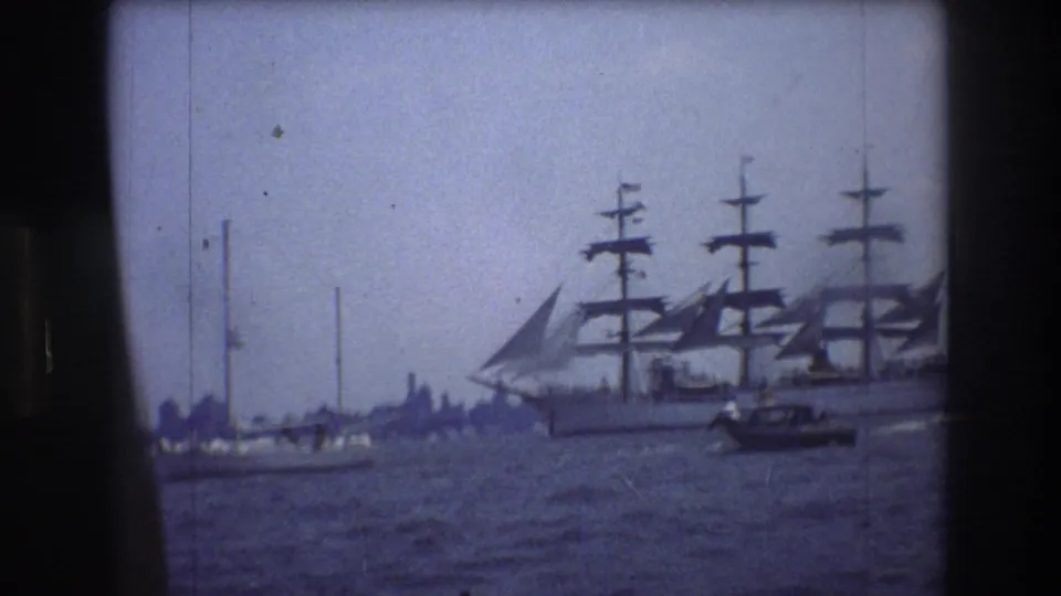 A large multi-masted sailing ship moves across New York City harbor water in 1976 home-movie footage, with smaller boats nearby and a hazy shoreline in the distance.