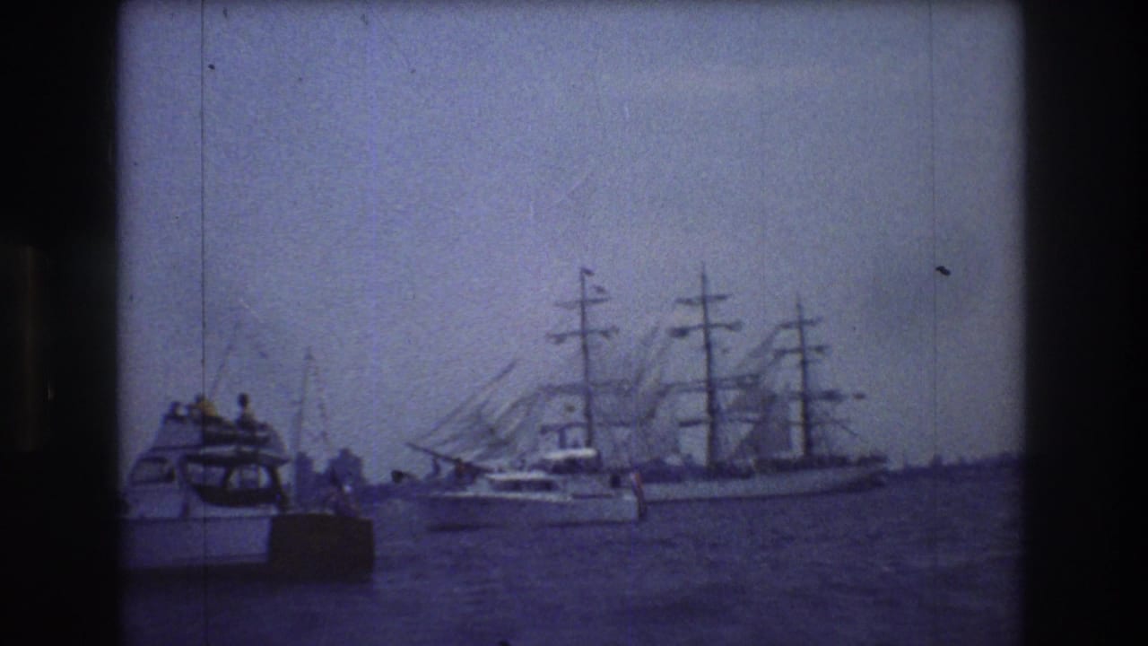 A small motorboat with people aboard sits in the foreground while a multi-masted sailing ship passes behind it on New York City harbor water.