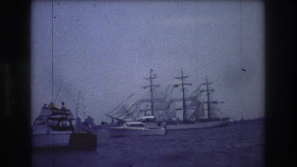 A small motorboat with people aboard sits in the foreground while a multi-masted sailing ship passes behind it on New York City harbor water.