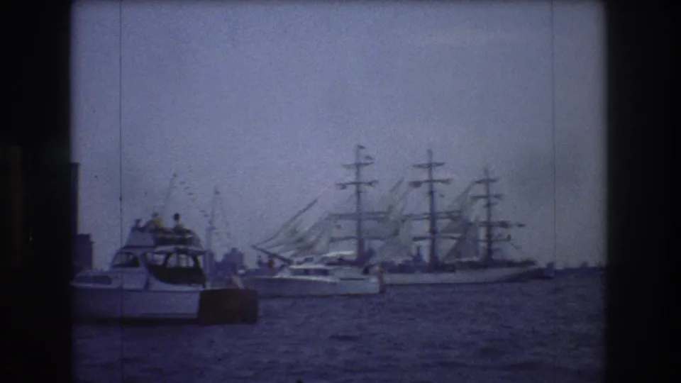 A small motorboat with people aboard crosses the foreground as a multi-masted sailing ship remains farther back on the hazy 1976 New York City harbor.