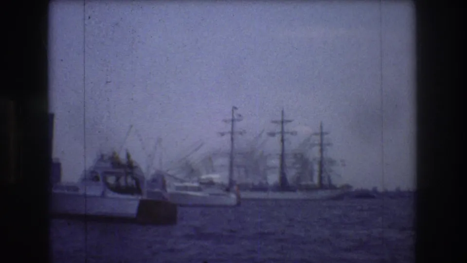 The sailing ship appears farther across the harbor with a small motorboat at left, softened by blue-tinted grain and haze in the 8mm home-movie frame.