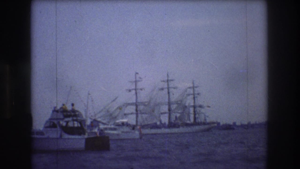 1976 New York City home-movie frame showing a small white motorboat with people aboard passing in front of a distant multi-masted sailing ship and harbor cranes.