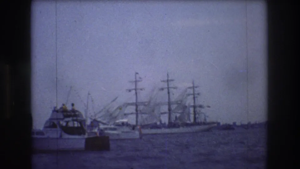 1976 New York City home-movie frame showing a small white motorboat with people aboard passing in front of a distant multi-masted sailing ship and harbor cranes.