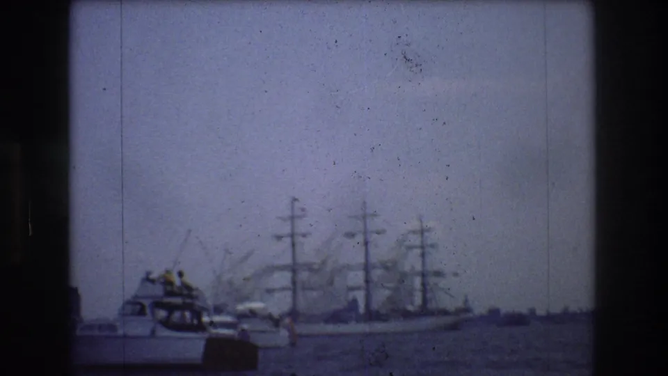 1976 New York City harbor view with a motorboat in the foreground, a multi-masted sailing vessel behind it, and industrial waterfront shapes fading into the haze.
