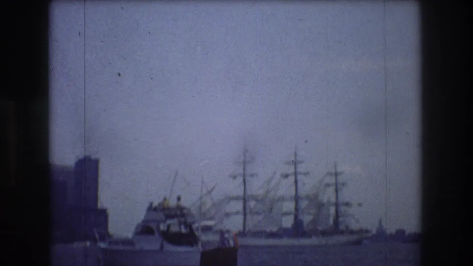 1976 New York City 8mm frame showing a motorboat crossing choppy water near a tall sailing ship, with blocky waterfront buildings and cranes in the distance.