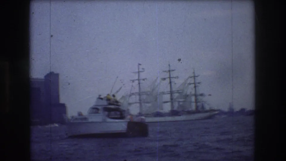 1976 New York City harbor frame with a white motorboat in the foreground, a multi-masted sailing ship behind it, and distant buildings under a pale hazy sky.