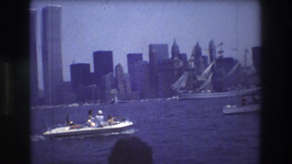 A motorboat with passengers crosses the harbor in front of Lower Manhattan, with the World Trade Center towers and other skyscrapers visible in the background.