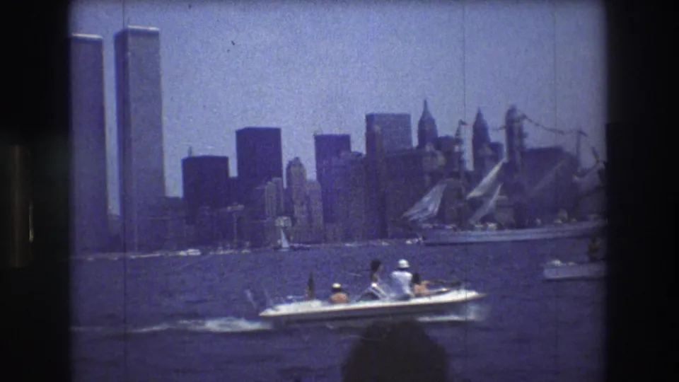 A small passenger motorboat travels across choppy harbor water with the World Trade Center towers, Manhattan skyline, and sailboats behind it.