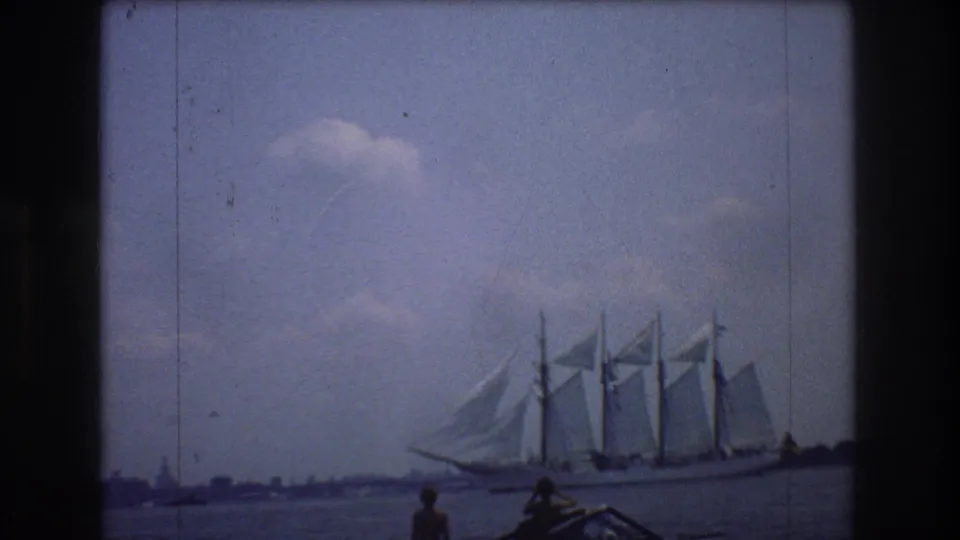 1976 8mm New York City harbor frame showing a multi-masted sailing ship under sail while two boating onlookers watch from the foreground.