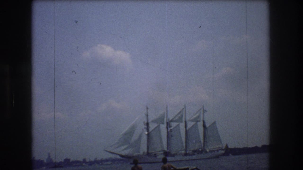 A multi-masted tall ship sails across New York harbor in 1976 footage, with a low shoreline and small distant spectators or passengers near the frame edge.