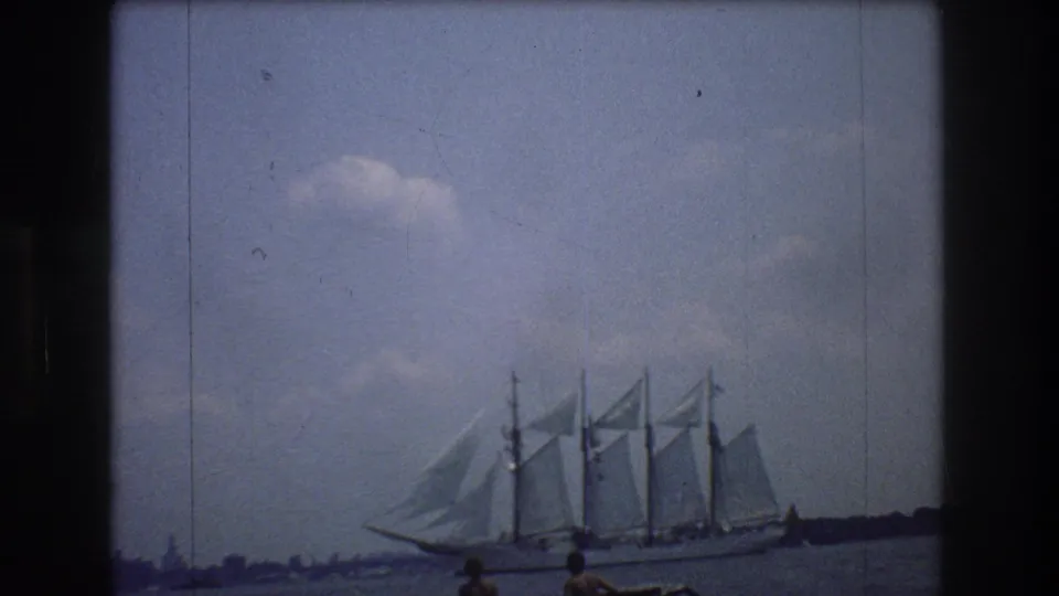 A multi-masted tall ship sails across New York harbor in 1976 footage, with a low shoreline and small distant spectators or passengers near the frame edge.