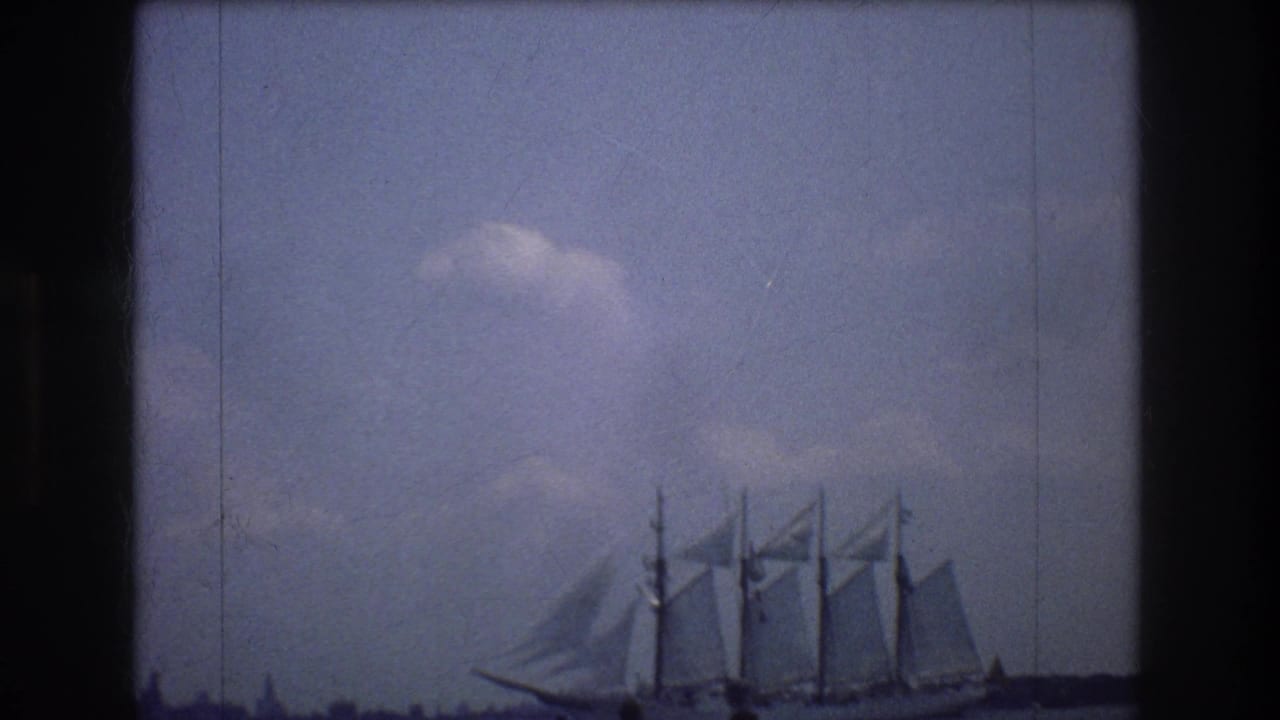 The tall ship remains in view on New York harbor, its masts and sails silhouetted softly against a blue, cloudy 1970s home-movie sky.