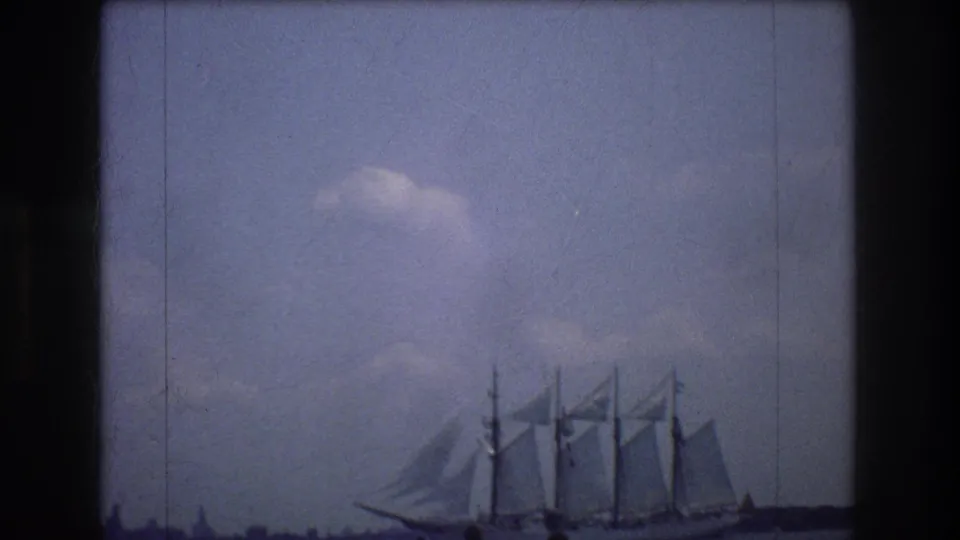 The tall ship remains in view on New York harbor, its masts and sails silhouetted softly against a blue, cloudy 1970s home-movie sky.