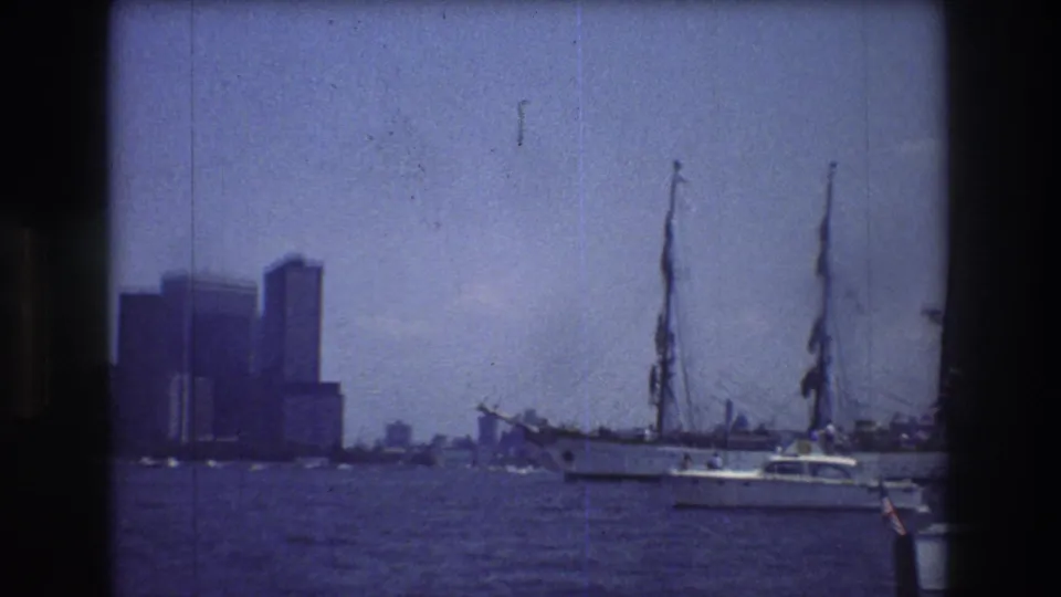 Manhattan buildings rise in the distance as sailboat masts and a small motorboat cross the harbor foreground in 1976 New York City footage.