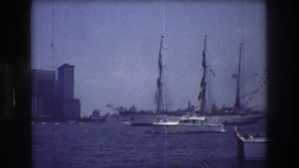 A small boat moves through choppy New York harbor water with sailboat masts, partial hulls, and distant Manhattan buildings visible behind it.