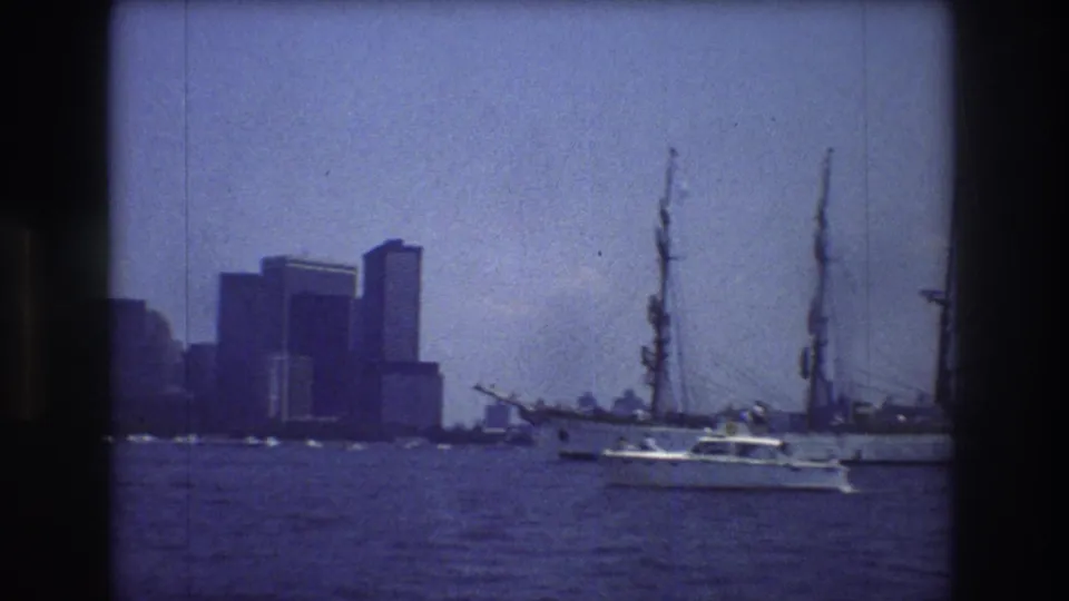 A small motorboat crosses beside a multi-masted sailing ship in New York City harbor, with 1970s Manhattan high-rises visible through haze.