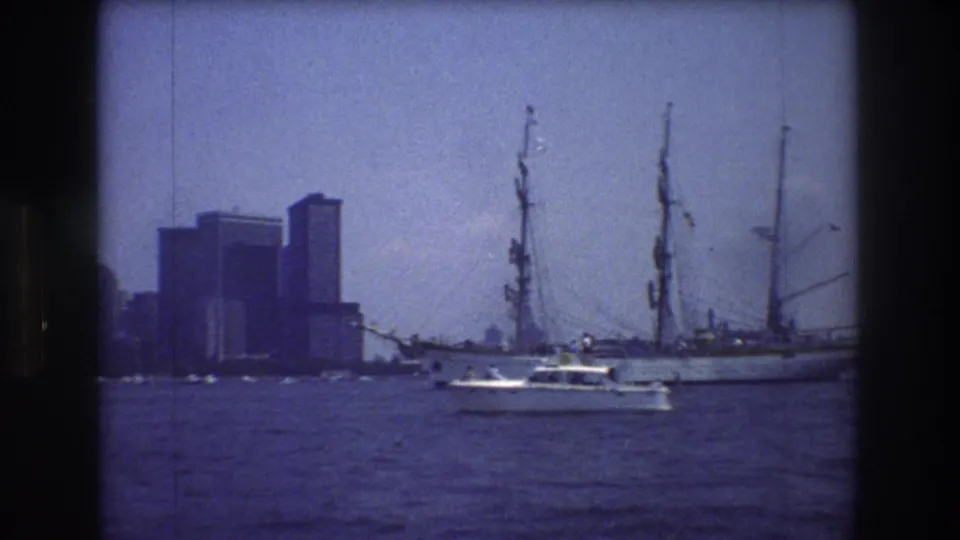 1976 New York harbor frame with a white motorboat in front of a large furled-sail vessel and blocky Manhattan skyline forms in the distance.