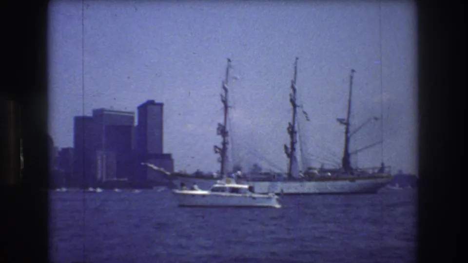1976 New York City harbor view showing a large white multi-masted sailing ship, a small motorboat crossing the foreground, and distant high-rise buildings under a blue cast.