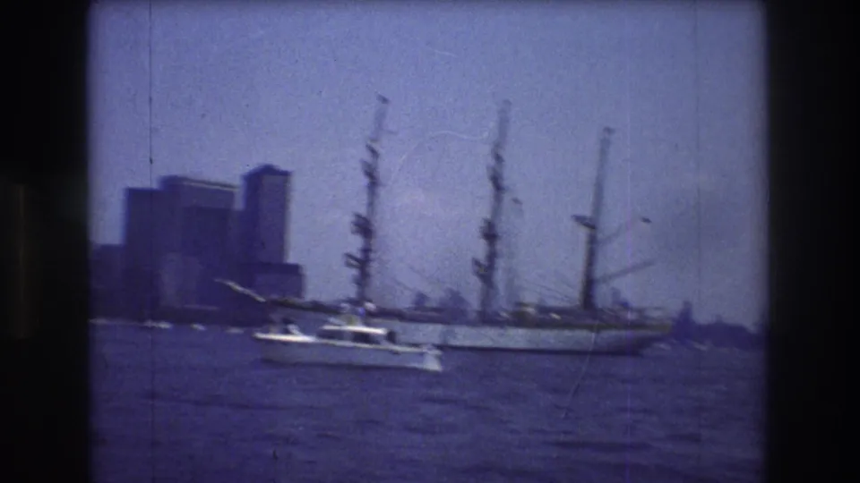 1976 New York City harbor frame with a small white motorboat moving across the water before a large multi-masted sailing ship and distant skyline.