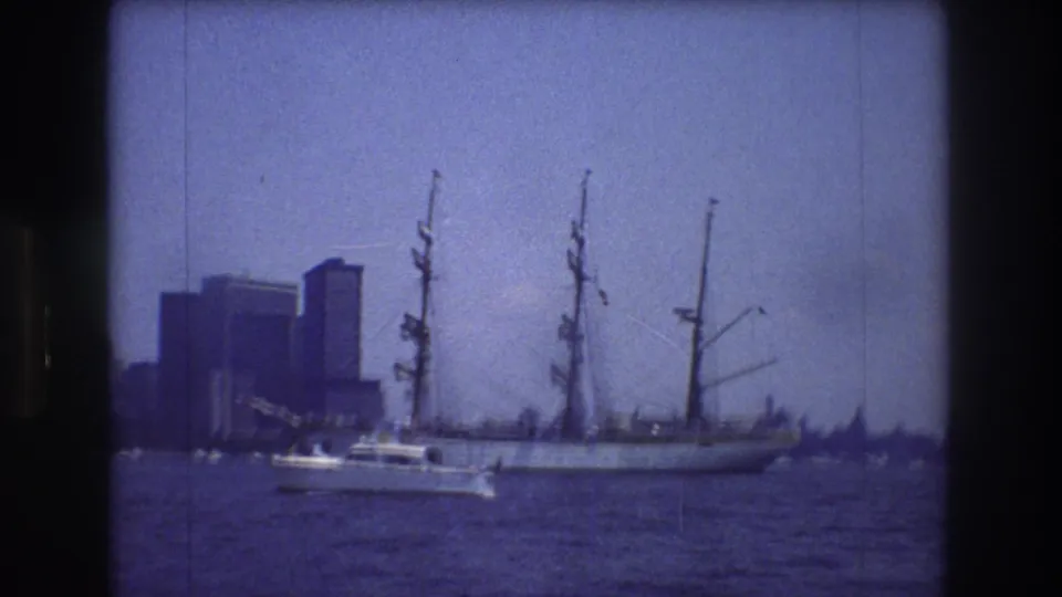 1976 New York City harbor view with the motorboat lower in frame, the large sailing ship partly centered, and dark high-rise buildings visible across the water.