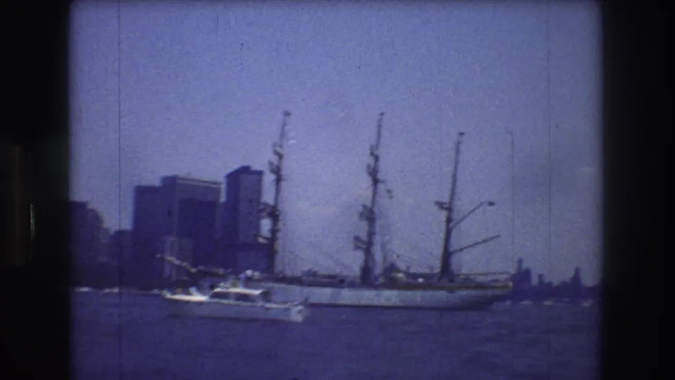 1976 New York City harbor frame showing a large white multi-masted sailing ship, a small motorboat near its side, and distant urban buildings through blue 8mm film color.