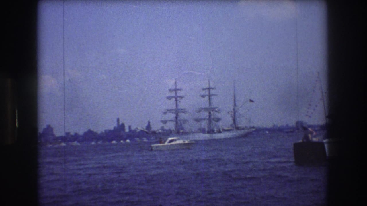 1976 New York City 8mm frame showing a large three-masted sailing ship moving across harbor water, with a small motorboat passing in front and faint shoreline shapes behind.