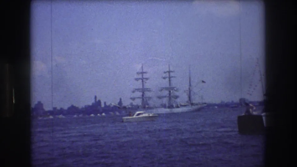 1976 New York City 8mm frame showing a large three-masted sailing ship moving across harbor water, with a small motorboat passing in front and faint shoreline shapes behind.