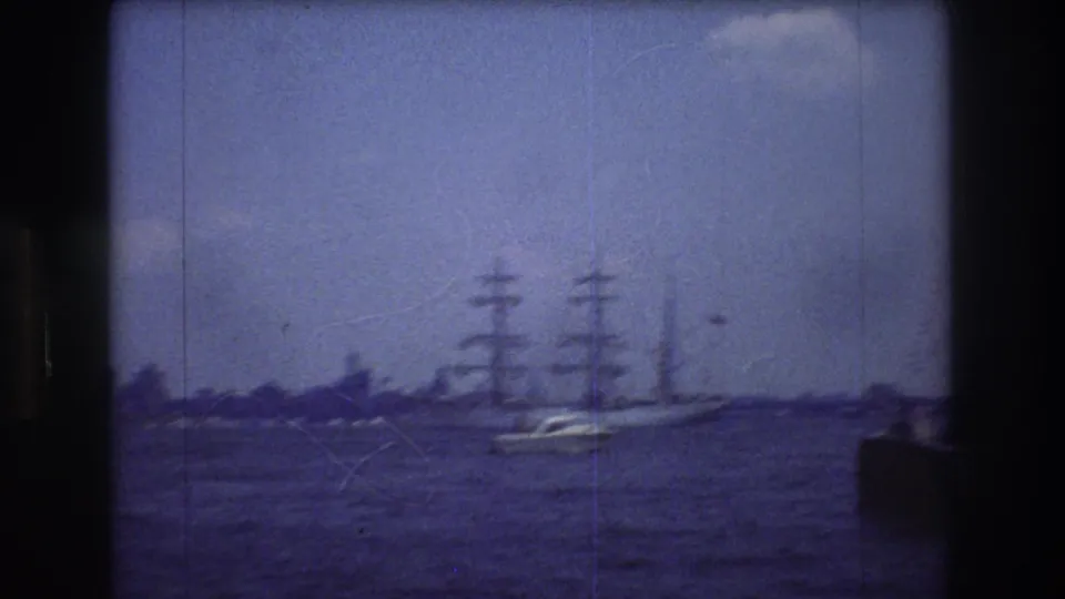 1976 New York City 8mm frame with a tall sailing ship seen at a distance on blue harbor water, faint shoreline behind it, and dark scan or camera-edge shadows at the sides.