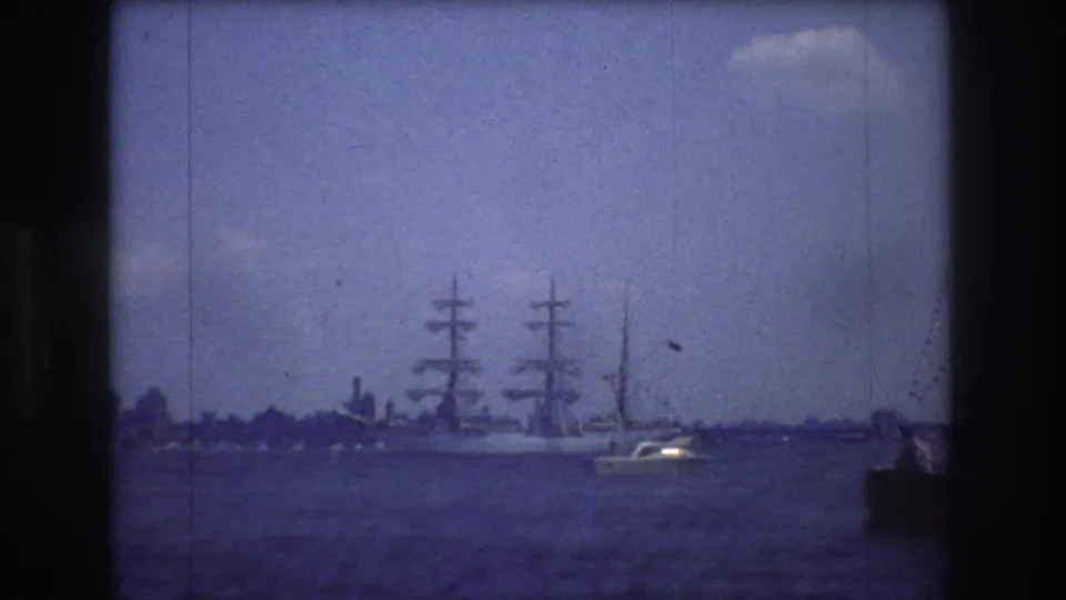 1976 New York City 8mm frame with the sailing ship near the horizon, a small motorboat crossing the foreground, faint city or shoreline forms in back, and visible film wear.