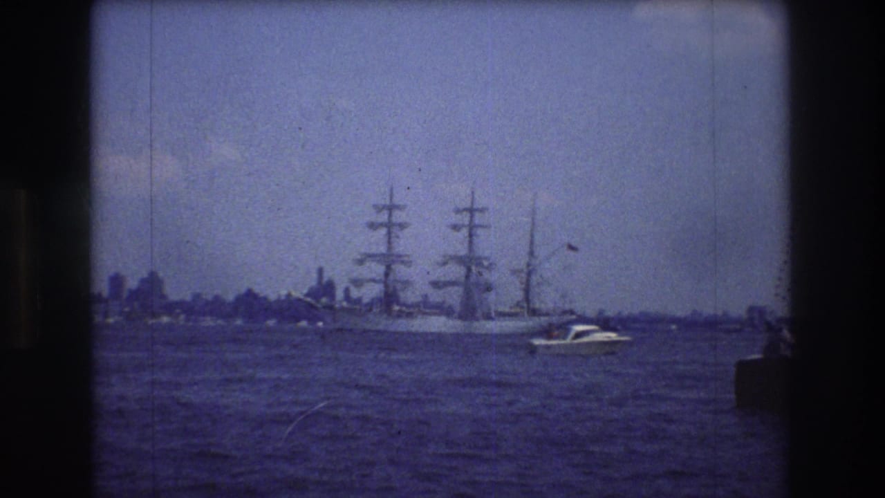 A 1976 8mm frame near New York City shows a large three-masted sailing vessel crossing open water, with a small motorboat in front and a hazy skyline behind.